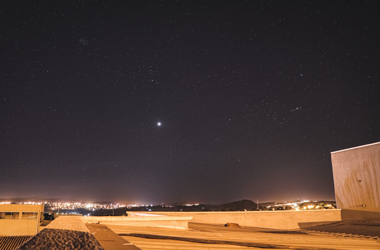 Ponto de luz muito brilhante no céu estrelado, visto do telhado de um prédio, com as luzes da cidade ao fundo